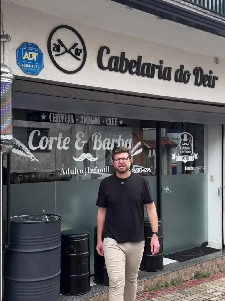 Deir em frente à fachada da Cabelaria do Deir, com a vitrine Corte & Barba, Adulto e Infantil