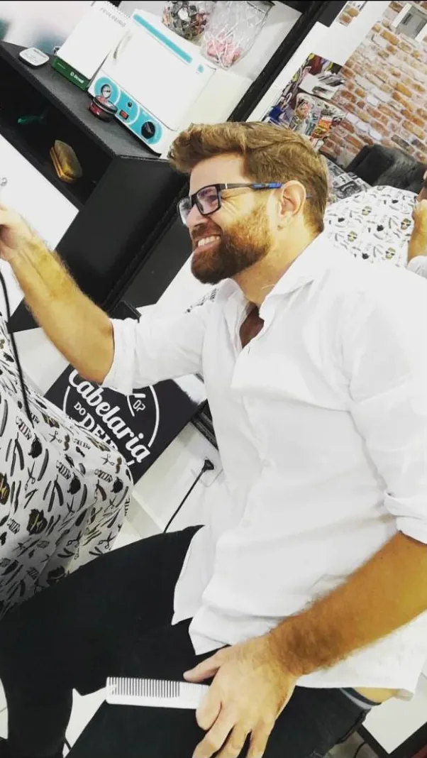 Deir sorrindo enquanto acerta a barba de um cliente com a máquina